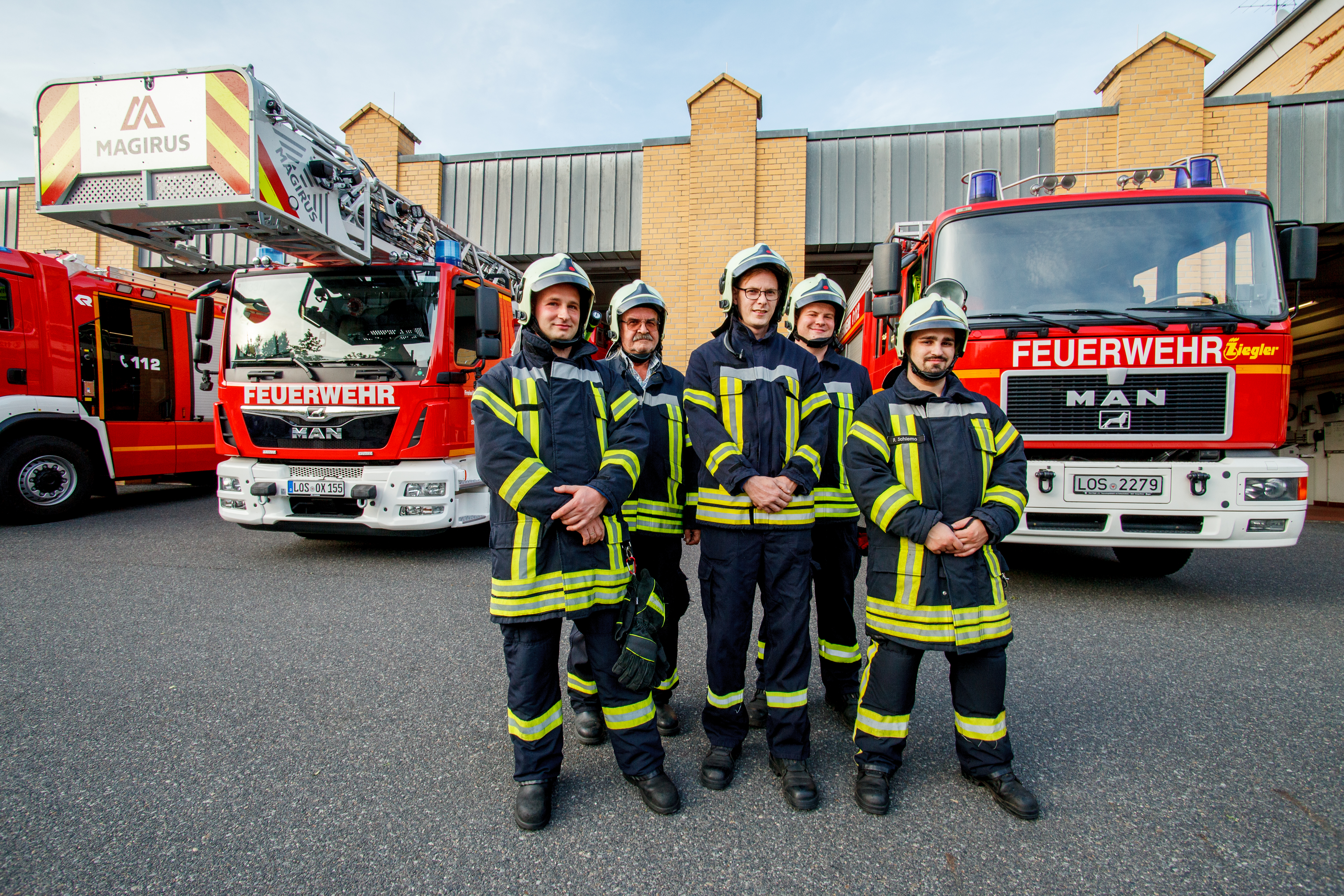 fünf Feuerwehrmänner posierend vor Einsatzwagen und Feuerwache